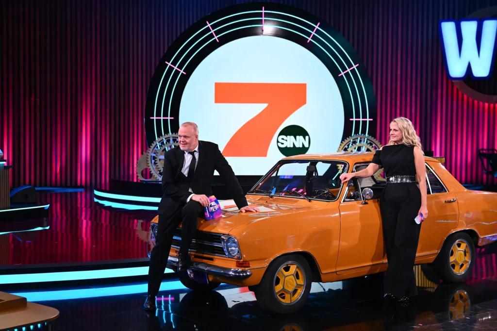 Moderationspaar mit einem orangefarbenen Oldtimer im Studio von „Wer weiß wie wann was war?“
