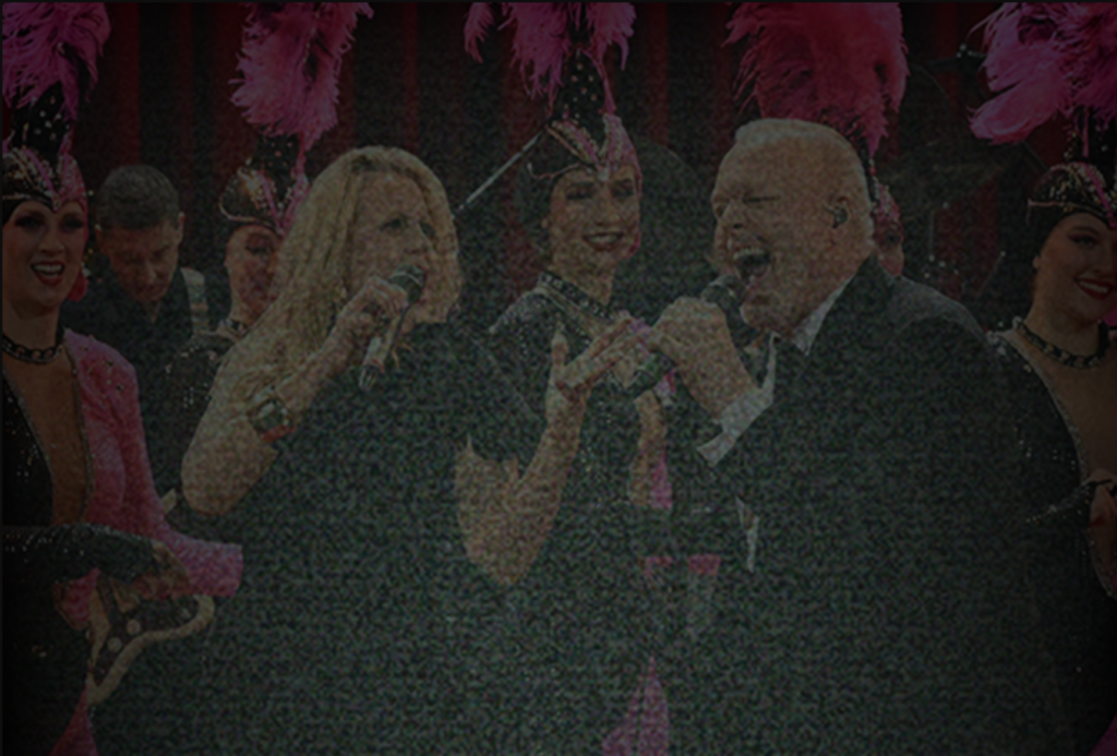 Bühnenszene: Sängerin und Sänger performen vor Showtänzerinnen mit Federschmuck